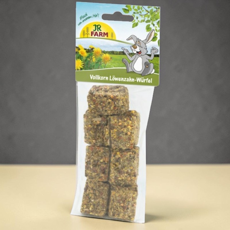 Transparent packaging containing seven square, brown pet treat cubes, filled with grains and herbs. Top card: a cheerful rabbit in a sunny field of dandelions, with the inscription ‘JR Farm Wholemeal Dandelion Cubes’.