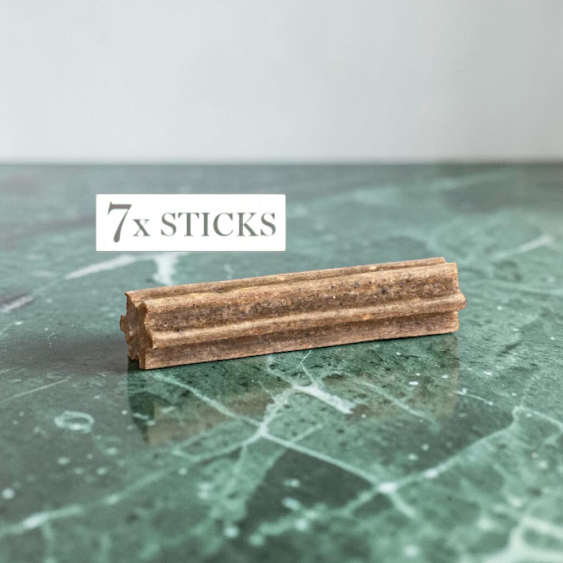 Un bâtonnet à mâcher allongé marron clair repose sur un plateau en marbre vert foncé brillant. Sa surface est striée de rainures longitudinales. Une étiquette blanche indique « 7x STICKS ».