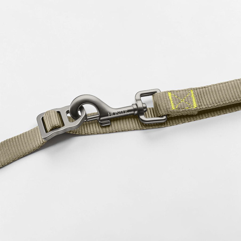 Detailaufnahme einer robusten graugrünen Hundeleine mit feiner Textur und gelbgrünen Ziernähten. Im Fokus: matter dunkler Alu-Sicherheitskarabiner mit Schnellverschluss, "HUNTER" graviert, am Verstellring.