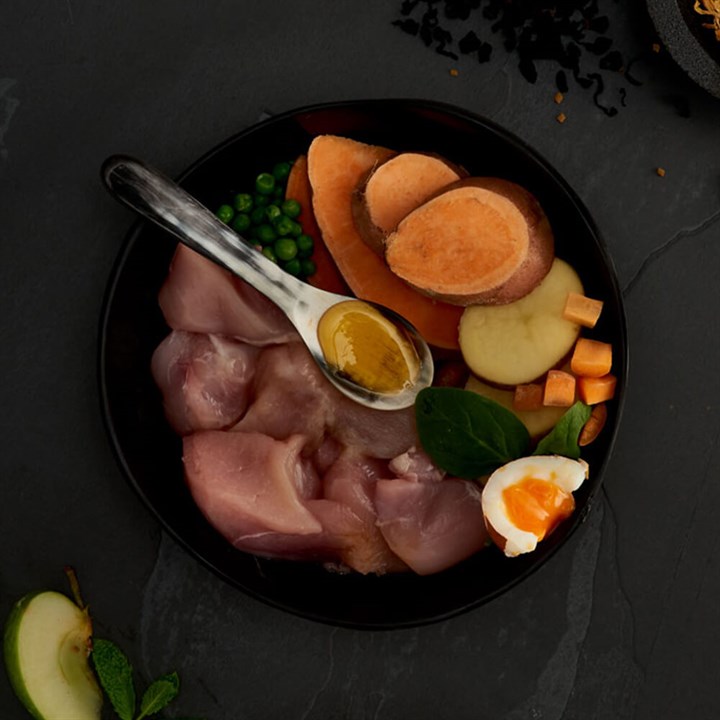 Dans une gamelle noire : morceaux de poulet cru élevé en plein air, rondelles de patate douce orange, petits pois verts, carottes en dés et demi-œuf dur. Une cuillère de liquide doré par-dessus. Aliments pour chiens Canagan.