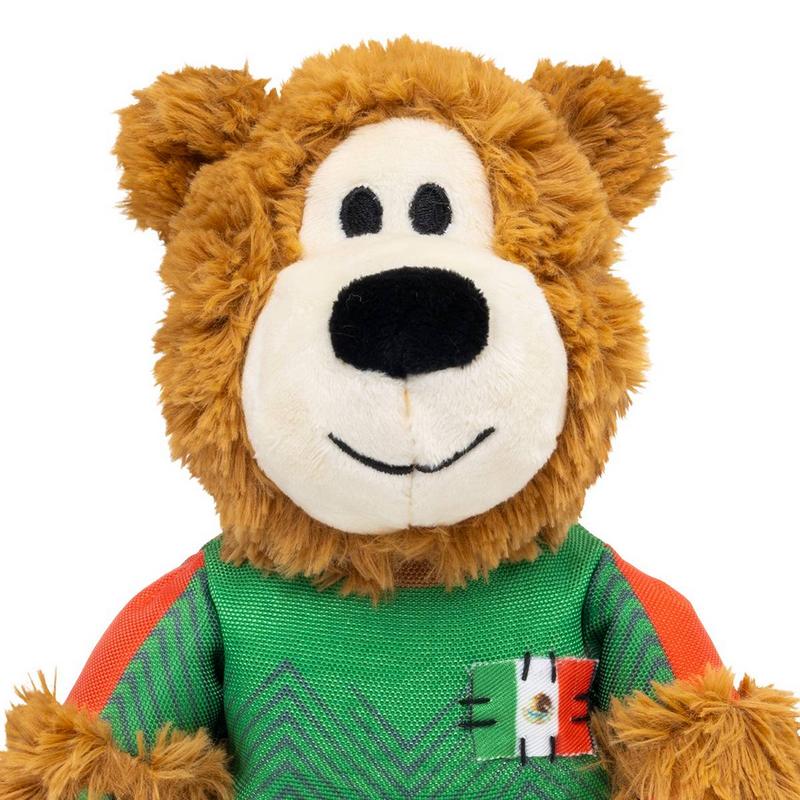 Un morbido orsacchiotto di peluche marrone guarda dritto verso la fotocamera. Indossa una maglietta verde con motivo a zigzag e maniche arancioni. Sul lato sinistro del petto è cucita una bandiera messicana.