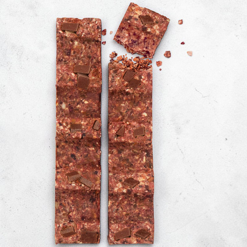 Due barrette di muesli marrone-rossastre, disposte verticalmente con pezzetti di cioccolato fondente, su sfondo grigio chiaro e ruvido. Quella a sinistra è integra; a destra manca un pezzo con briciole.