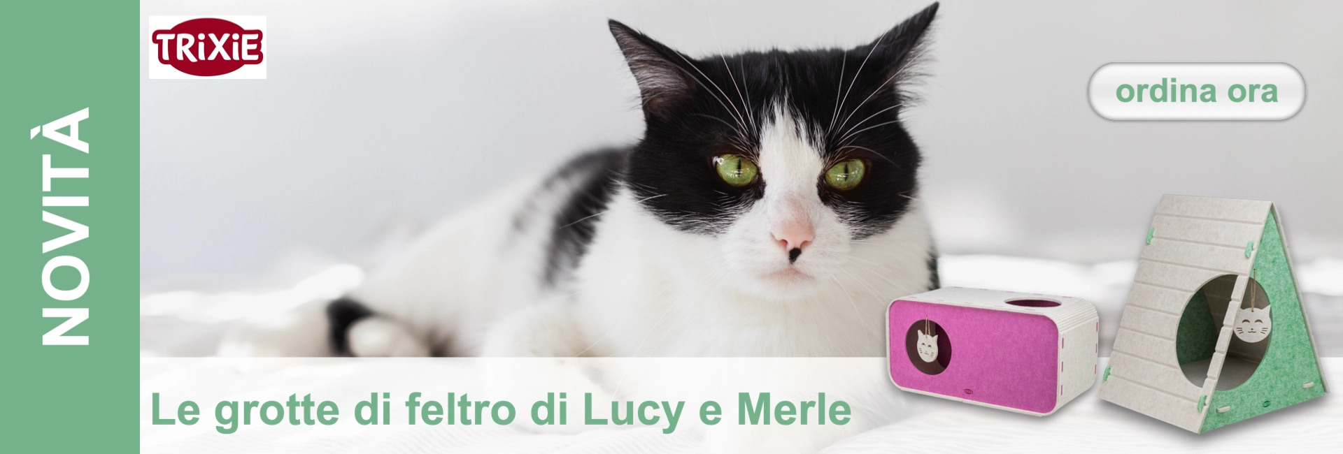 Gatto bianco e nero con intensi occhi verdi giace su uno sfondo chiaro e guarda verso la fotocamera. In primo piano due cucce in feltro: una rosa, rettangolare, e una verde, triangolare, con ingressi rotondi e faccine di gatto.