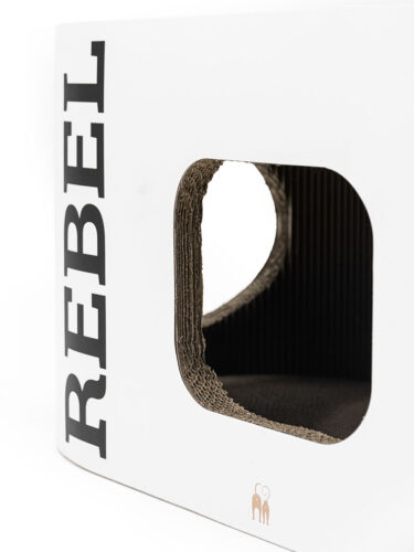 Tana in cartone bianco con apertura rettangolare arrotondata sul davanti, che mostra l'interno in cartone ondulato marrone e uno spazio buio. A sinistra è riportata la scritta "REBEL" in verticale in nero. In basso a destra un piccolo logo con due gatti.