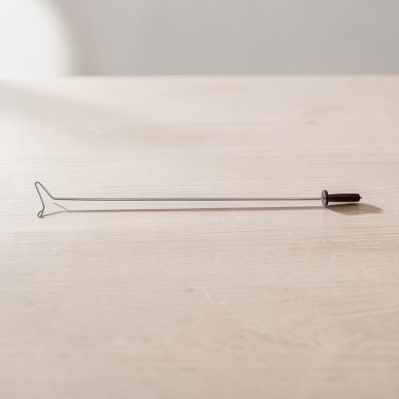 Ein langer, schlanker Metallstab liegt auf einer hellen Holzfläche. Links ist ein kleiner, nach oben gebogener Haken, rechts ein kurzer, dunkler, zylindrischer Griff. Das Werkzeug ist parallel zum unteren Bildrand.