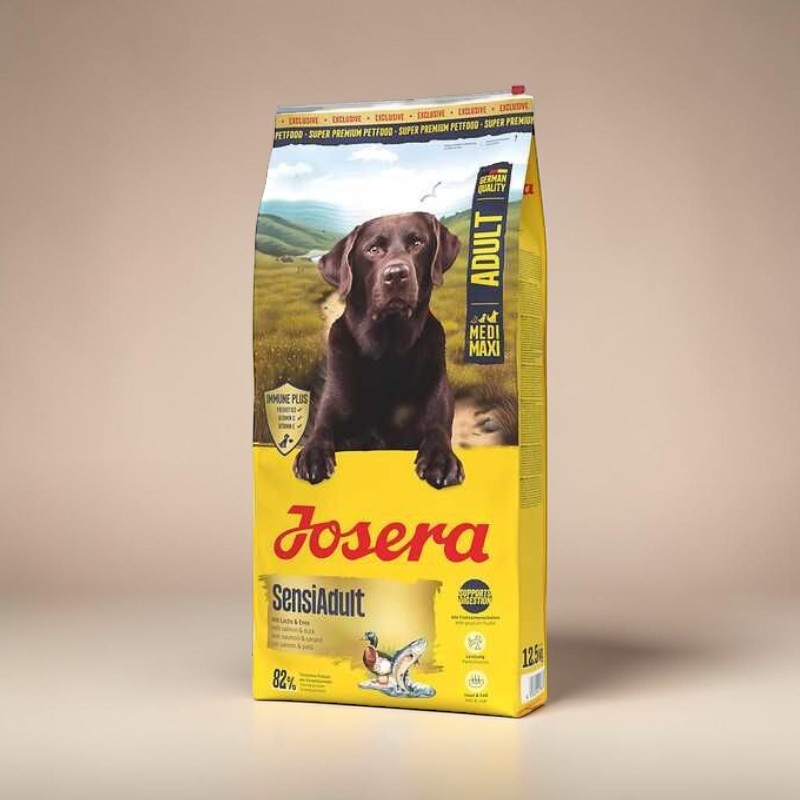 Confezione gialla del mangime per cani SensiAdult di Josera, destinato a cani adulti. Un labrador marrone guarda in avanti, con le zampe appoggiate sulla busta. Sfondo: colline verdi, cielo blu. Scritte: IMMUNE PLUS, MEDI MAXI, GERMAN QUALITY.