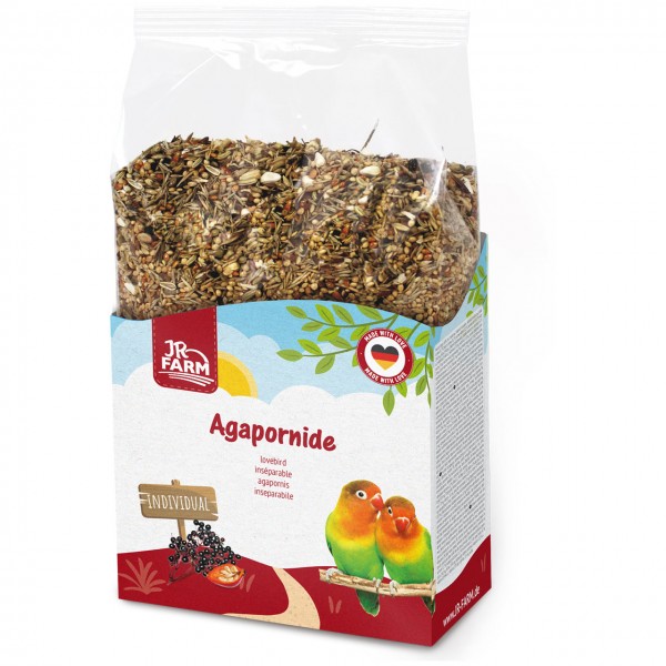 Bag of mixed bird feed for lovebirds in colourful cardboard packaging. Featuring the JR Farm logo, “Agapornide” and the Made with love seal. Two green lovebirds with orange heads on a branch. Wooden sign “INDIVIDUAL” and dark berries.