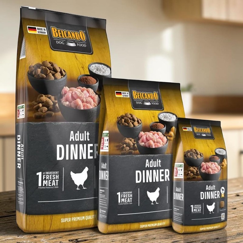 Tre sacchetti dorati e neri di croccantini per cani "Belcando" Adult Dinner, in diverse confezioni, su una superficie in legno. Nell'immagine: croccantini, carne di pollame e polvere. In evidenza: "Adult DINNER" e icona del pollo.