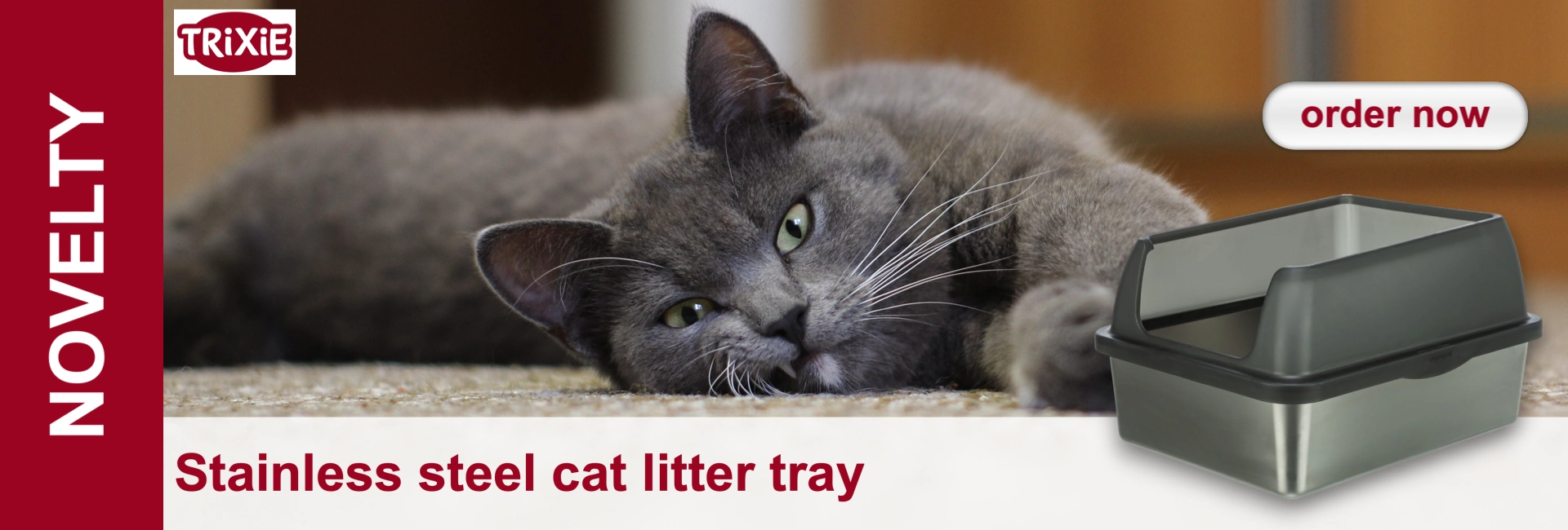 A relaxed grey cat is lying on a light-coloured carpet, looking straight into the camera; its green eyes have heart-shaped pupils. To the right of it is a shiny stainless steel litter tray with a dark grey lid.