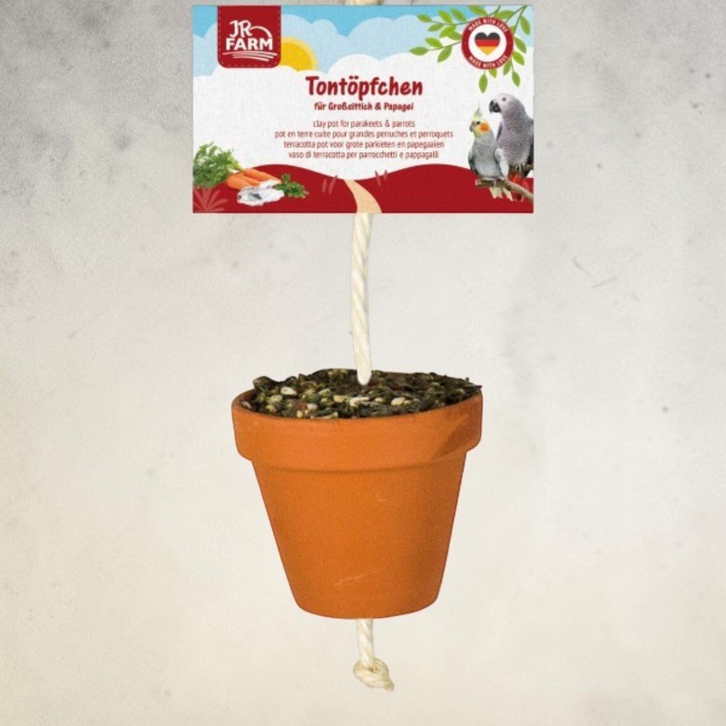 A small brown terracotta pot containing a birdseed mix hangs from a white rope. Above it is a white label reading ‘JR Farm Terracotta Pot for Large Parakeets & Parrots’, along with pictures of vegetables and two parrots.