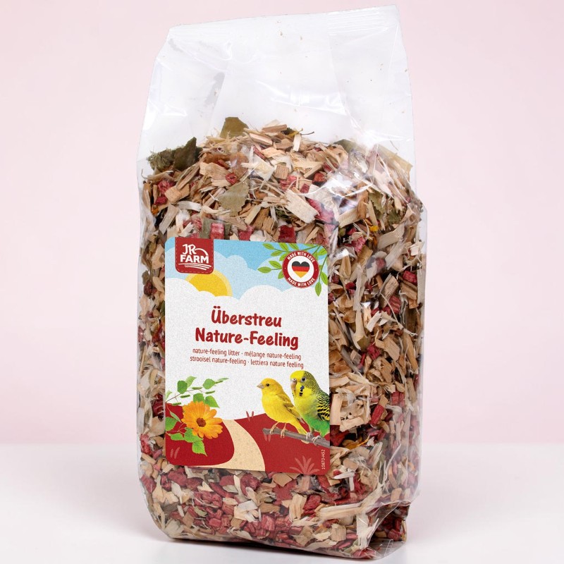 Durchsichtige Plastiktüte mit JR FARM Überstreu Nature-Feeling für Kleintiere, gefüllt mit Holzspänen, getrockneten Blättern und roten Pflanzenmaterialien. Etikett: Firmenlogo, Kanarienvogel und Wellensittich auf Ast, orange Ringelblume.
