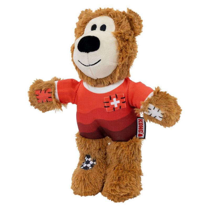 Morbido peluche marrone a forma di orso per cani. Indossa una maglietta a righe rosse con la croce svizzera sul petto e una toppa da calcio sul piede sinistro. Faccia simpatica con muso bianco e occhi neri. Etichetta Kong sul braccio.