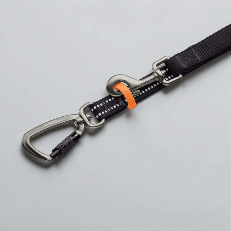 Gros plan sur la laisse Hunter Action ErgoMove Pro noire, avec points blancs réfléchissants. Équipée de deux mousquetons argentés : un mousqueton de sécurité en aluminium à fermeture rapide et clip, plus un amortisseur orange.