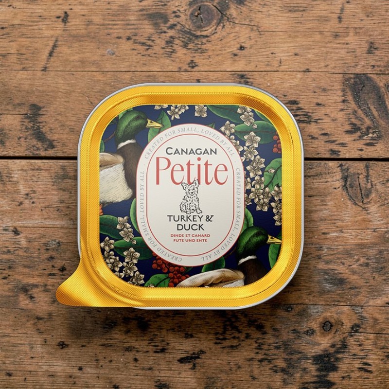 Barquette alu jaune « Canagan Petite » de nourriture humide pour chiens, sur bois rustique. Couvercle : scène bleu foncé avec canards, fleurs blanches et baies rouges. Texte : « Canagan Petite Turkey & Duck ».