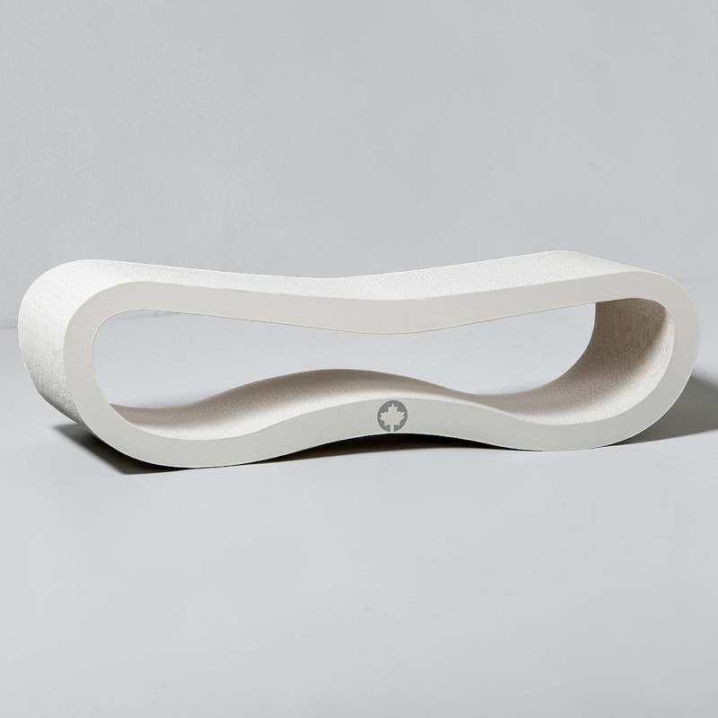 An elongated, white object with a gently curved, organic shape rests on a light grey surface. In the centre is a wave-shaped opening. The matt surface has a fine texture. At the bottom centre is a dark leaf logo.