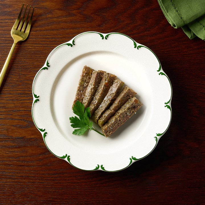 Tranches prêtes à servir d’une délicieuse galette d’agneau pour petits chiens, joliment présentées sur une assiette blanche à motifs verts. On voit la texture grossière de la nourriture humide, avec une branche de persil. Fourchette dorée à côté.