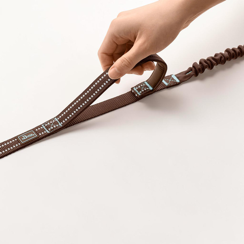 Eine Hand hält eine braune Hunter Action-Hundeleine mit reflektierenden weißen Punkten. Die ErgoMove Pro Leine hat eine gepolsterte Schlaufe und einen Ruckdämpfer gegen plötzliche Zugbewegungen. Das hellblaue Hunter-Logo ist sichtbar.