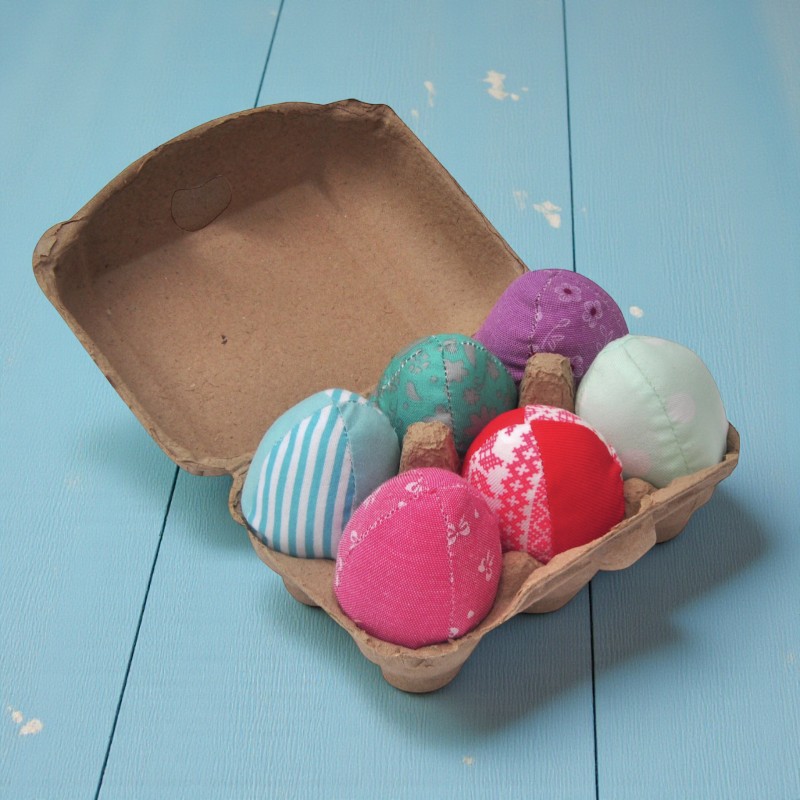 Sechs bunte Stoff-Ostereier für Katzen liegen in einem geöffneten Eierkarton auf blauem Holzboden.