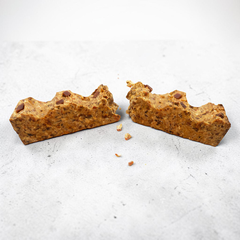 Barretta rettangolare marrone dorato spezzata al centro su superficie grigio chiaro. Ha consistenza granulosa con pezzetti scuri simili a gocce di cioccolato. Briciole sparse vicino alla rottura.