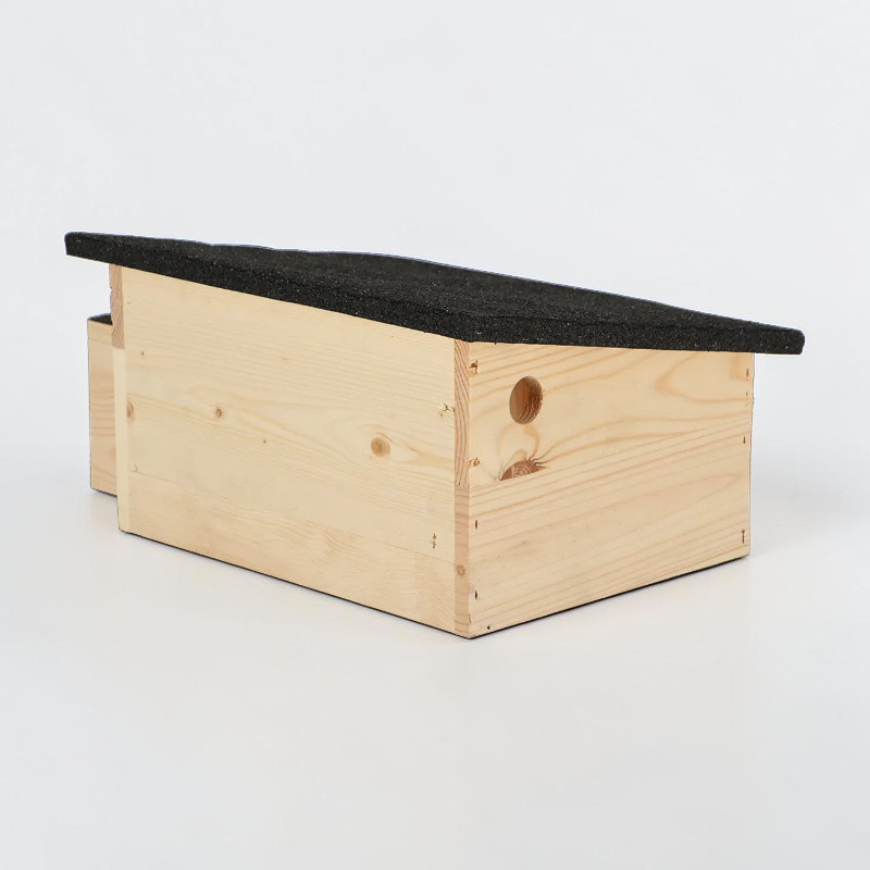 Abri pour hérissons L en bois d'épicéa clair, avec toit incliné recouvert de bitume foncé. Abri allongé en bois avec ouverture d'aération ronde sur le côté et tunnel d'entrée visible sur le côté court, refuge sûr.