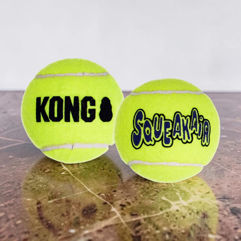 Zwei leuchtend neongelbe Tennisbälle liegen auf einer glänzenden braunen Oberfläche. Der linke Ball trägt das schwarze Logo "KONG", der rechte Ball ist mit "SqueakAir" in Blau und schwarzem Umriss beschriftet. Der Hintergrund ist hell und unscharf.