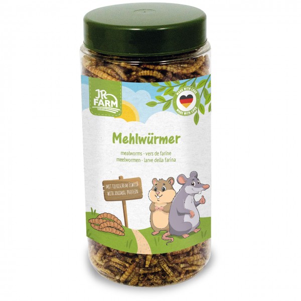 Contenitore trasparente con coperchio verde, riempito di larve della farina essiccate. Etichetta: "JR Farm", "Larve della farina", illustrazione con criceto e topo accanto al cartello "CON PROTEINE ANIMALI". Cielo soleggiato sullo sfondo.