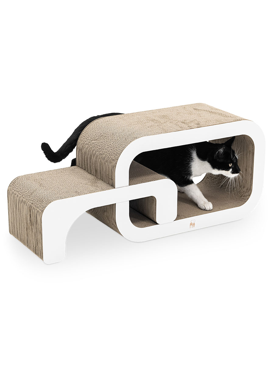 Schwarz-weißer Kater kriecht neugierig aus einem modernen Kratzmöbel aus beiger Wellpappe mit weißen, abgerundeten Rahmen. Kopf und Vorderpfoten ragen aus dem Tunnel, der Schwanz hinten heraus. Blick nach rechts.