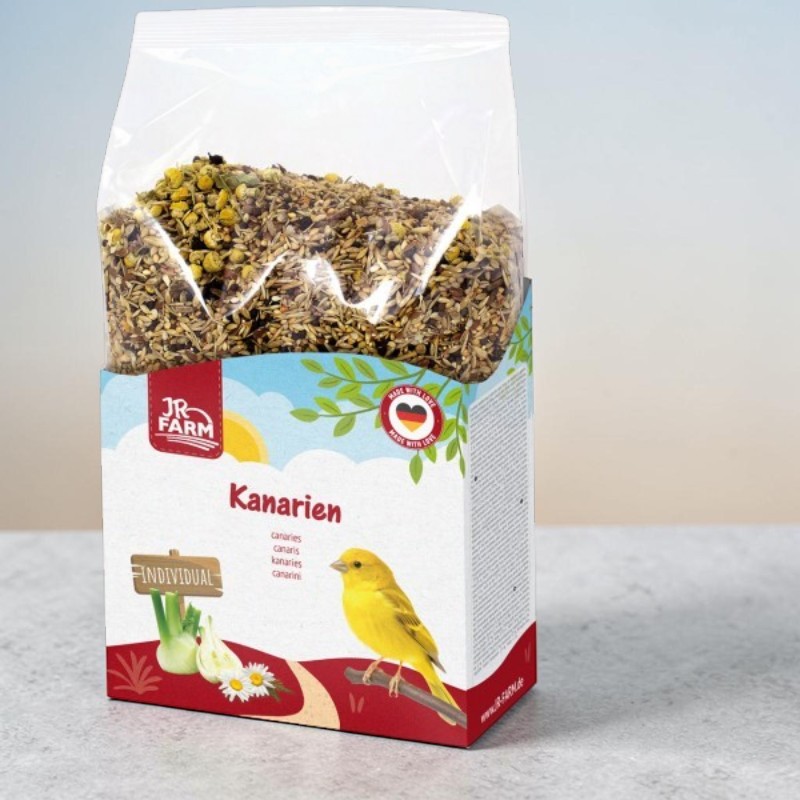 Sachet transparent de nourriture pour canaris JR Farm contenant des graines, des céréales et des fleurs de camomille séchées. Étiquette colorée représentant un canari jaune, du fenouil et des fleurs blanches. Inscriptions « Kanarien » et « Individual ».
