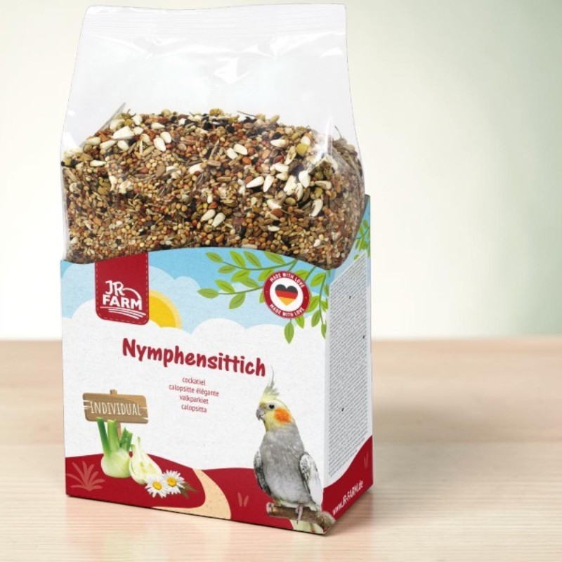 Emballage transparent de nourriture pour perruches calopsittes à base de graines et céréales. Banderole en carton colorée avec la marque « JR Farm », symbole de la calopsitte, fenouil et pâquerettes. «Nymphensittich» et «Made with Love» visibles.