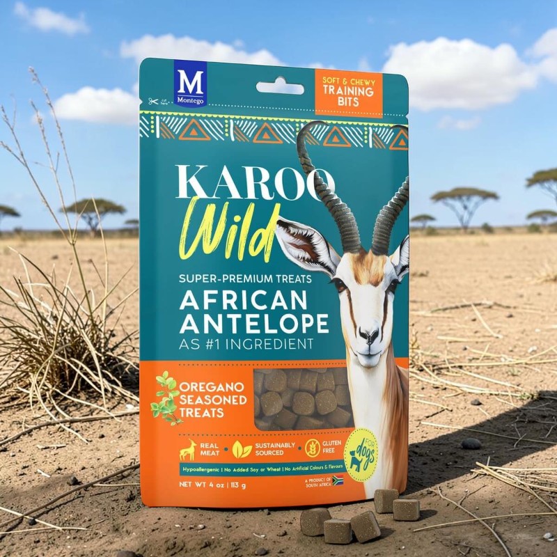 Confezione verde turchese e arancione con scritta "Karoo Wild" e "African Antelope" sul terreno secco della savana. Testa di springbok, "Oregano Seasoned Treats". Davanti cubetti. Sfondo: erba, alberi, cielo blu.