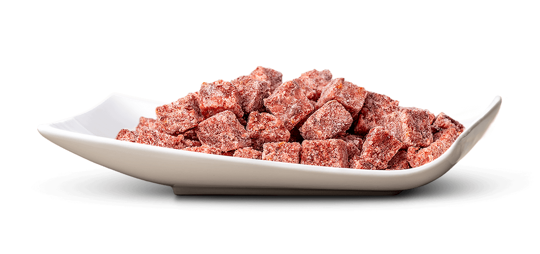 Nahaufnahme: Ein Berg kleiner, würfelförmiger, tiefroter Süßigkeiten, dicht mit feinem weißem Zucker bestreut, wirkt leicht frostig. Sie liegen in einer eleganten, länglichen, weißen Keramikschale auf reinweißem Hintergrund.