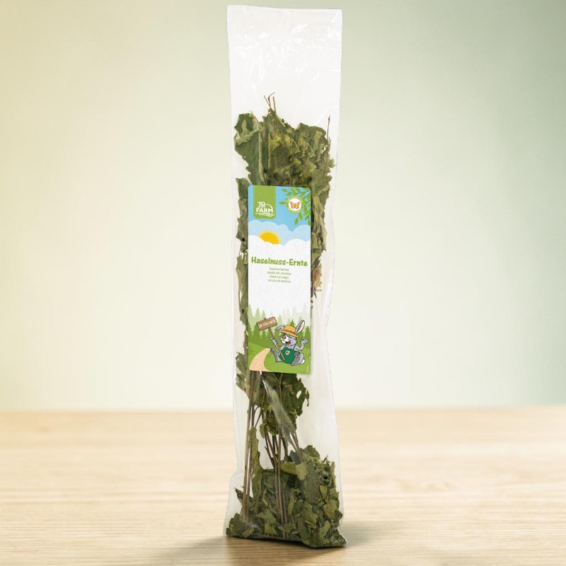 A transparent plastic bag containing dried hazelnut leaves and twigs is sitting on a light-coloured wooden surface. The bag has a coloured label bearing the words ‘Hazelnut Harvest’, a cheerful rabbit in work clothes and the JR Farm logo.