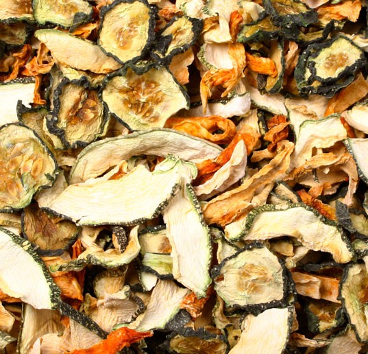 Close-up of a mixture of dried vegetable pieces: light green, round courgette slices with dark edges, as well as irregular orange and cream-coloured pieces, similar to carrots and root vegetables. Dry, slightly wavy.