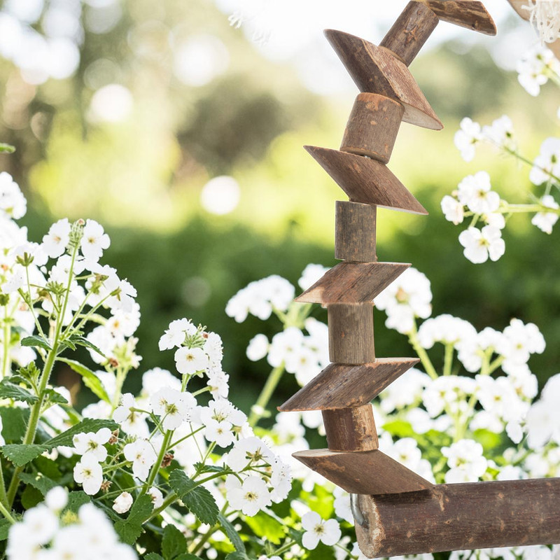 Gros plan d'une balançoire en bois naturel pour oiseaux, faite de morceaux d'écorce cylindriques et en coin, empilés verticalement et inclinés à droite. Bois rugueux, arrière-plan flou avec fleurs blanches et feuilles vertes.