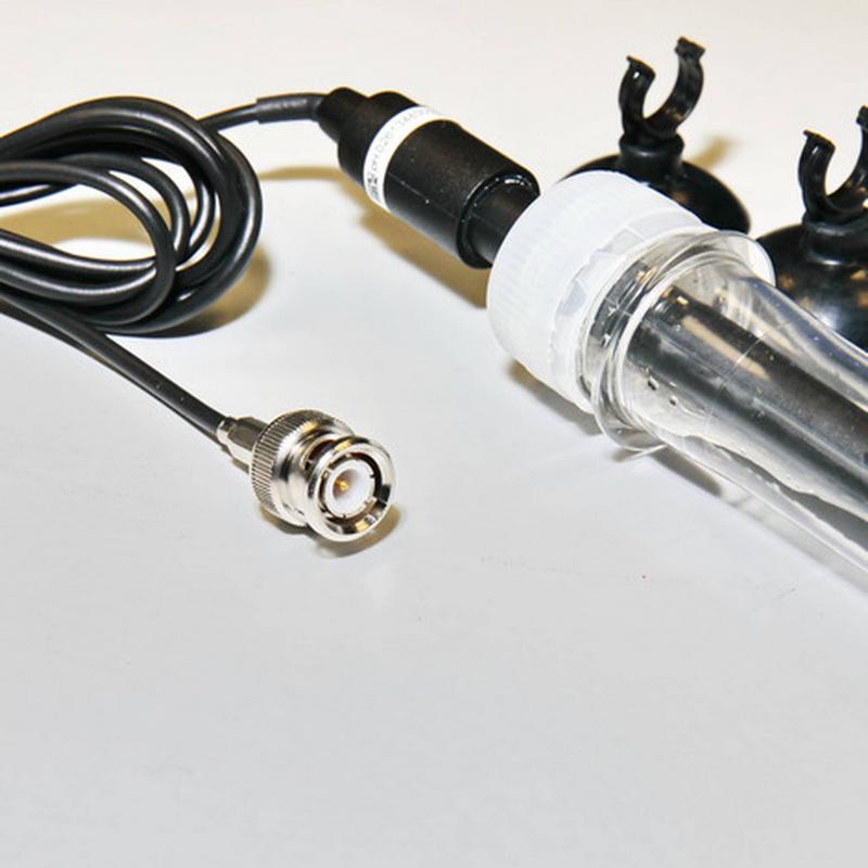 Gros plan sur un câble noir avec une fiche BNC brillante à gauche. Le câble mène à un composant cylindrique noir, relié à un tube transparent muni d'un embout blanc. À l'arrière-plan, deux ventouses noires.