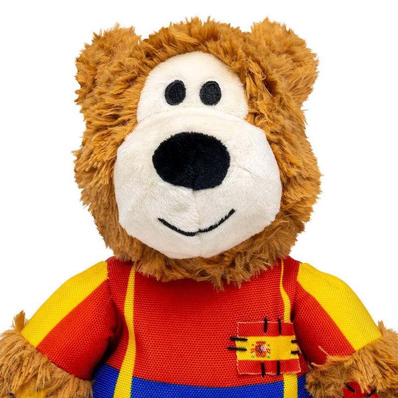 Un morbido peluche marrone con il viso chiaro, gli occhi e il naso neri, che sorride leggermente. Indossa una camicia colorata nei toni del rosso, giallo e blu. Sul petto c'è un piccolo distintivo con la bandiera spagnola e lo stemma.