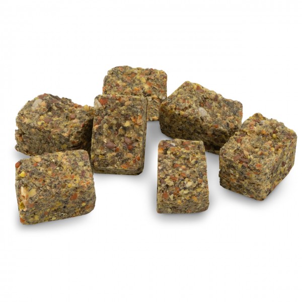 Eight irregularly shaped blocks made of pressed seeds and grains lie on a white background. They are greenish-brown with yellow, orange and reddish grains, look like bird or rodent feed, and have a rough, grainy texture.