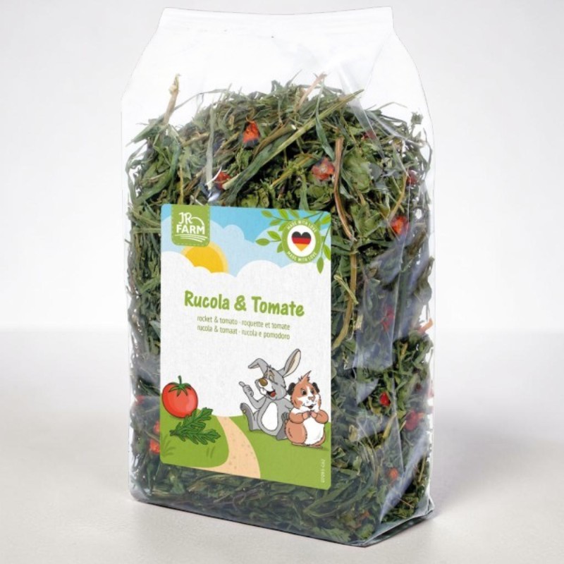 A transparent bag filled with dried green herbs and pieces of red tomato, as rodent food from the JR Farm brand. The label bears the words ‘Rocket & Tomato’, framed by a rabbit, a guinea pig, a tomato and rocket leaves.