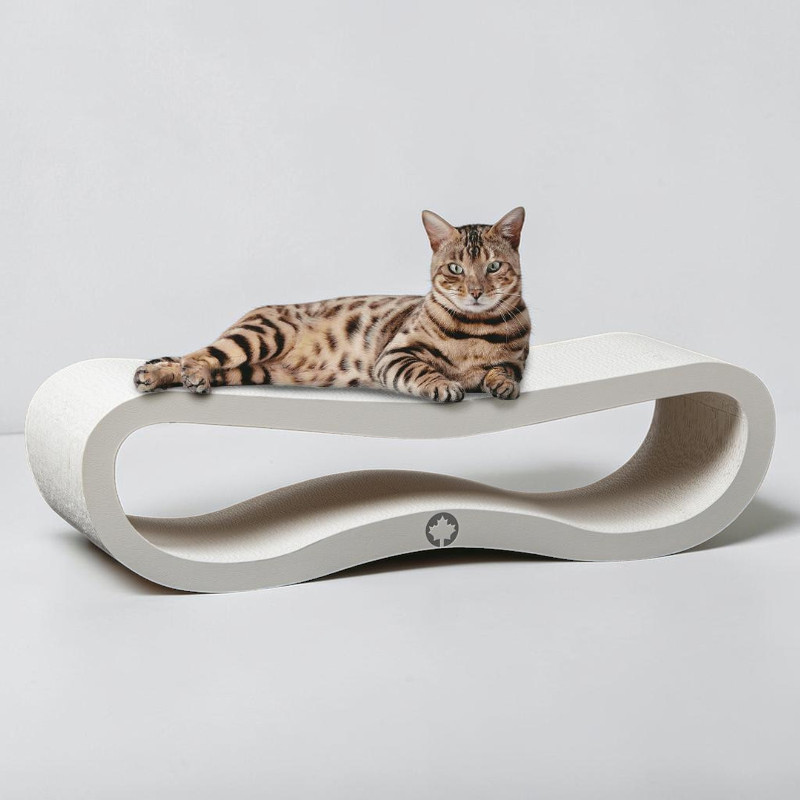 A Bengal cat with a tabby and spotted coat is relaxing on a modern, wave-shaped cat scratching post in light grey. It is looking at the camera with its green eyes whilst resting on the raised sleeping platform. The background is light.