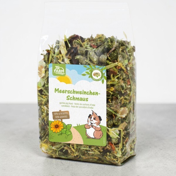 Transparent bag containing JR Farm Guinea Pig Feast, a dry supplementary feed made from green herbs, leaves and dried vegetable pieces. Label: Product name, guinea pig in a meadow, sunflower; ‘100% Pure Nature’, ‘Grain-free’.