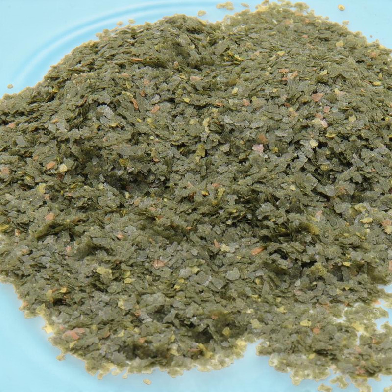 Feine, grüne Futterflocken für Medaka/Japanische Reisfische liegen als kleiner Haufen auf hellblauer Oberfläche. Unregelmäßige Flocken mit orange-braunen Partikeln, z.B. Spirulina, Lachs, Garnelen. XS für kleine Mäuler.