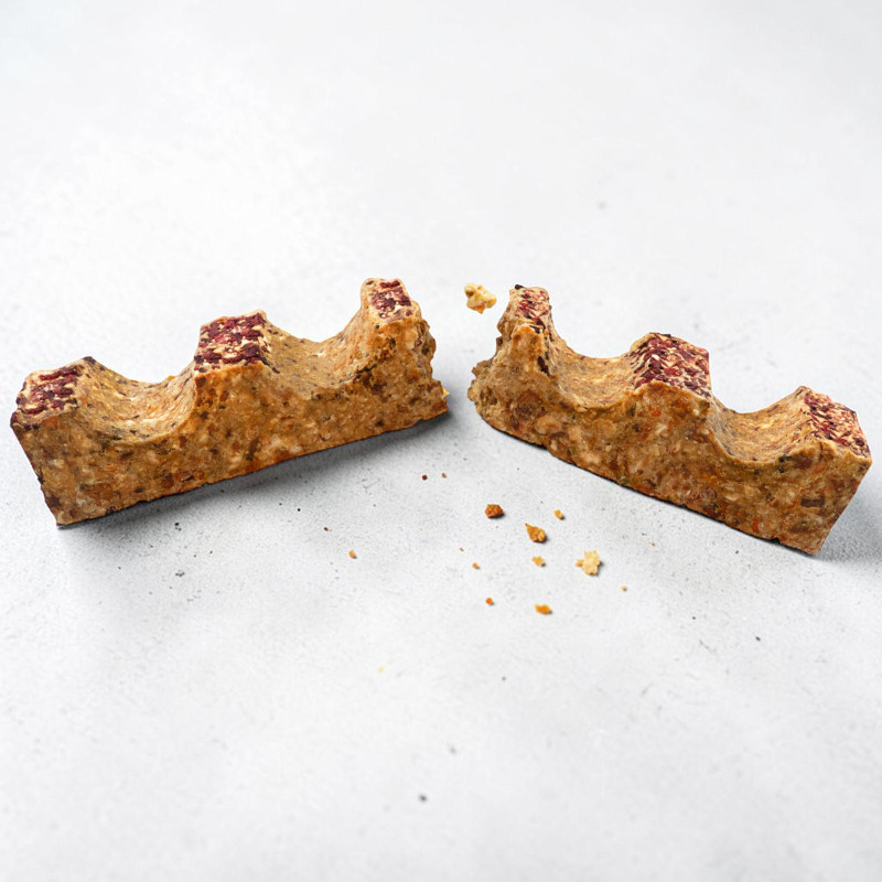 Barretta di muesli spezzata, consistenza irregolare e superficie ondulata tipo mura di castello con granelli rossastri. Cereali e noci marrone chiaro, rotta al centro con interno ruvido. Briciole su sfondo grigio chiaro.