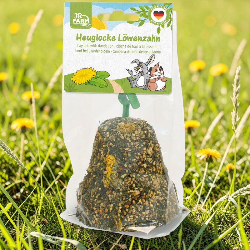Packaged JR Farm hay bell made from dandelions in a green meadow with dandelion flowers. Bell-shaped treat made from hay, seeds and dried dandelions. Label featuring the brand and product name, as well as a rabbit and a guinea pig.