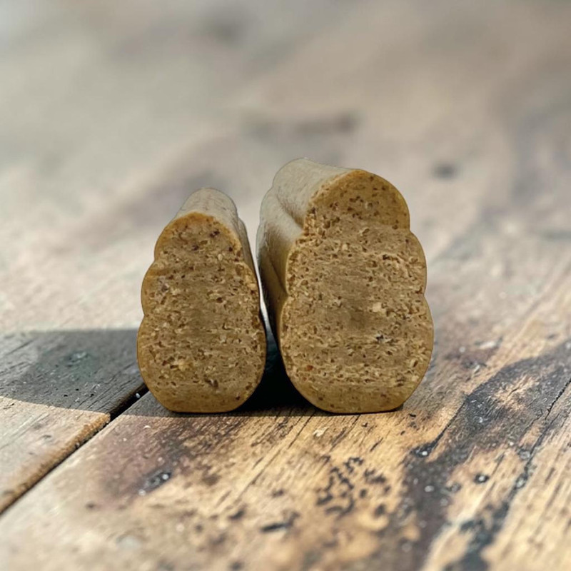 Zwei längliche, hellbraune Kauartikel mit rillenartiger, strukturierter Oberfläche stehen aufrecht auf einem rustikalen Holztisch. Links der kleinere, rechts der größere. Körnige Textur mit dunklen Sprenkeln, leichte Schatten.