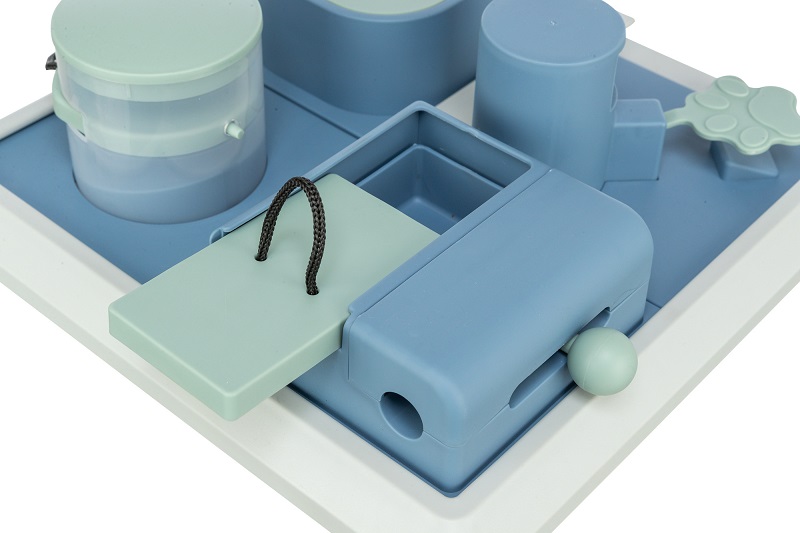 Jouet interactif pour animaux aux couleurs pastel, avec distributeur de friandises transparent, volet vert menthe avec cordelette au-dessus d’un récipient bleu, curseur à bille et levier en forme de patte sur plaque bleu clair.