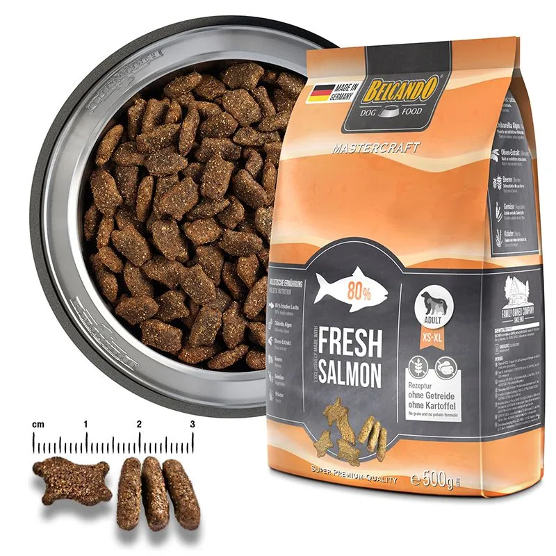 Orange and black bag of ‘ Belcando ’ Mastercraft Fresh Salmon dry food next to a filled metal bowl. Advertised as containing 80% fresh salmon, grain-free and potato-free. Three dark brown kibbles (one paw-shaped, two oblong) with a scale, approx. 1–2 cm.