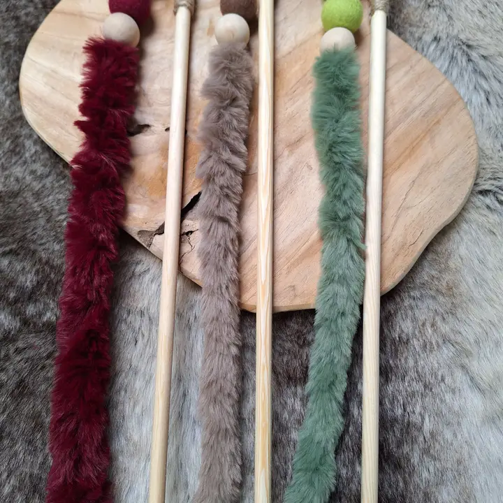 Drei Stäbe mit farbigen Fellstreifen und Kugeln, liegen auf einem Holzbrett vor einem Fellteppich.