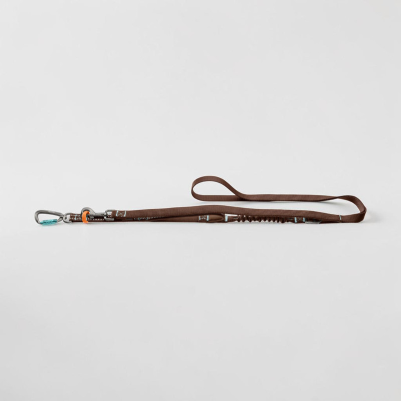 Braune Action-Hundeleine, flach auf hellem Untergrund, mit grauem Aluminium-Sicherheitskarabiner und blauen Akzenten links. Mit Reflexelementen, integriertem Ruckdämpfer und gepolsterter Handschlaufe rechts.