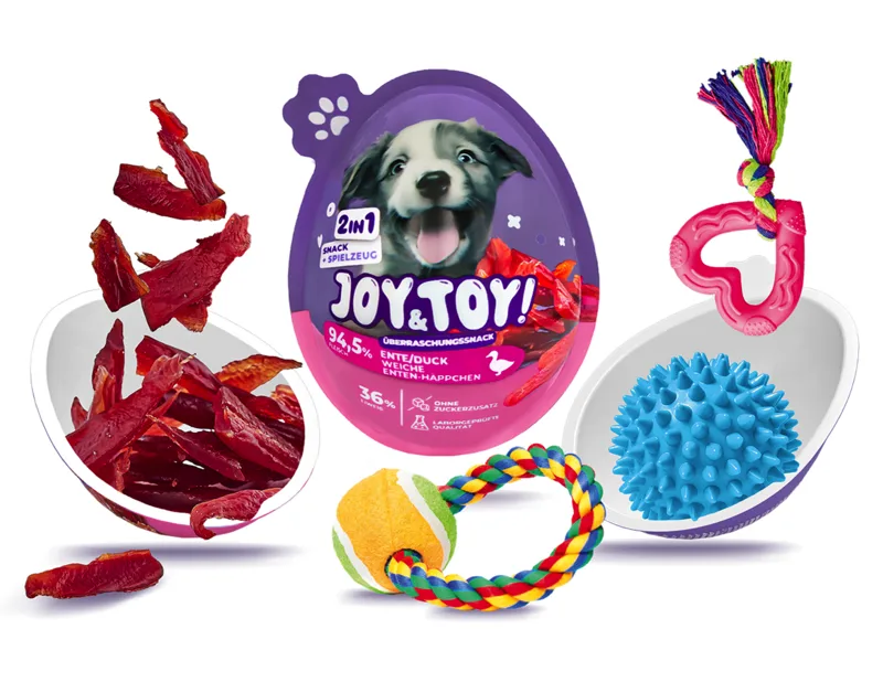 Weißer Hintergrund: lila Überraschungsei „Joy & Toy“ für Hunde mit lachendem Welpen. Links rote Entenfleischstreifen aus weißer Schale. Rechts blaue Igelkugel in Schale und pinkes herzförmiges Kauspielzeug. Vorn Tennisball-Seil-Spielzeug.
