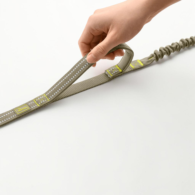 Eine Hand hält eine grau-grüne Action-Hundeleine mit reflektierenden Elementen und gepolsterter Handschlaufe. Rechts ist der integrierte Ruckdämpfer der ErgoMove Pro zu sehen, der Stöße abfedert. Hunter-Logo sichtbar.